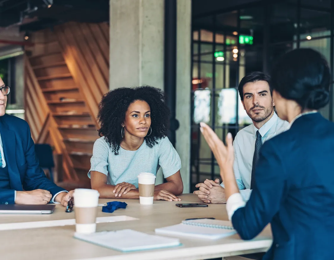 A group of five people in business attire sit around a table in a modern office setting. Four are facing the camera, listening attentively, while one, with their back to the camera, gestures as if speaking. Coffee cups and notebooks are on the table.