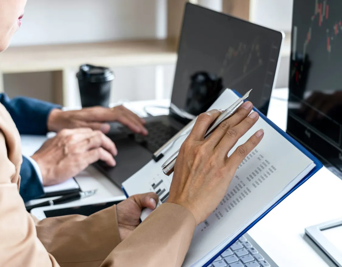 Two people working at a desk with laptops. One person is holding a pen and a document, while the other types on a laptop. A monitor displays a line graph with red and yellow lines. A coffee cup is in the background.