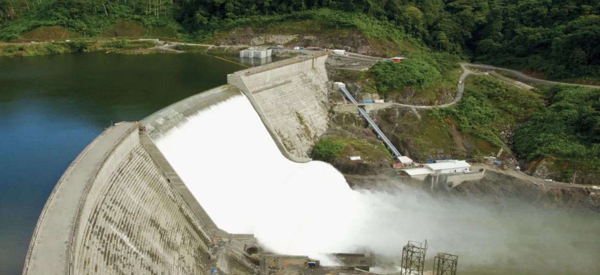 Aerial view of a large dam with water cascading over its spillway, surrounded by dense green forest. The dam structure is concrete, with water flowing into a river below. Roads and small buildings are visible near the dam, along with mist rising from the water.