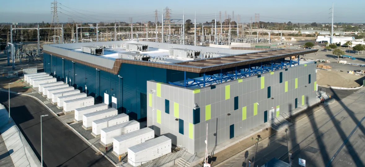 Aerial view of a large industrial facility with a modern, rectangular building featuring blue and gray panels with green accents. The building is surrounded by numerous white utility boxes and power lines. The area is paved with roads and pathways, and there are trees and other industrial structures in the background.