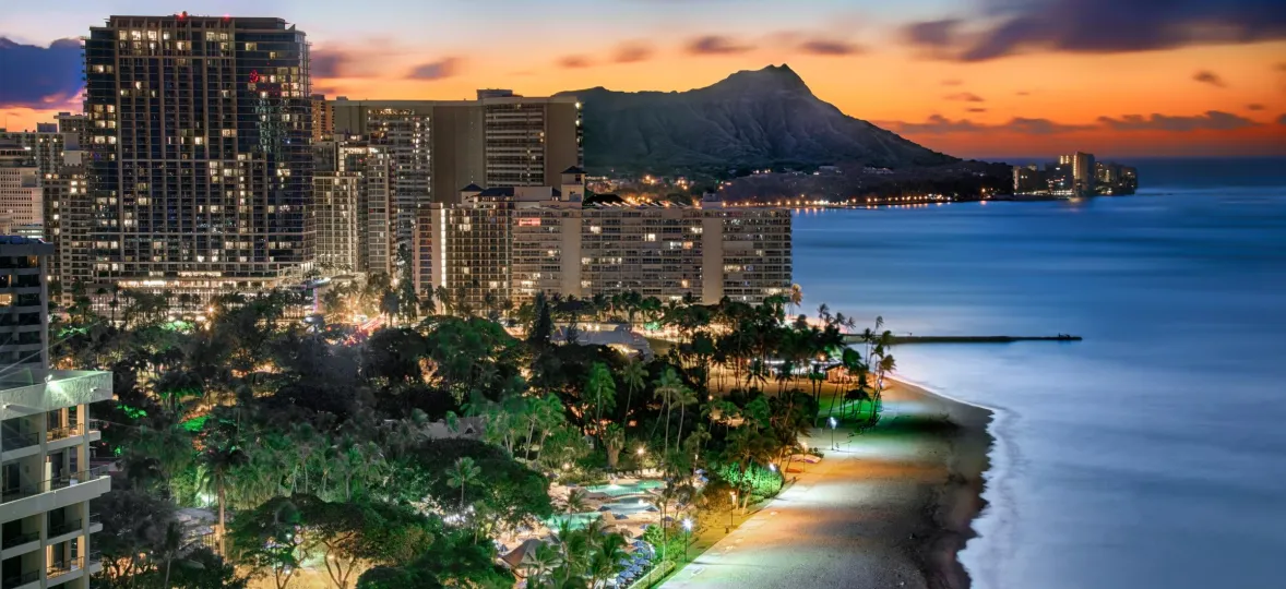 A cityscape at dusk with illuminated buildings near a beach. The ocean is calm, and a mountain is visible in the background under a vibrant sunset sky.