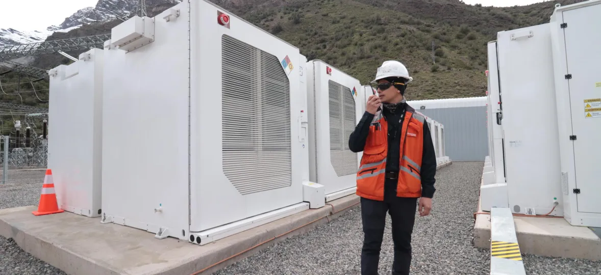 A person wearing a hard hat and orange safety vest holds a radio while standing in front of large white industrial units at a power facility. There are mountains in the background.