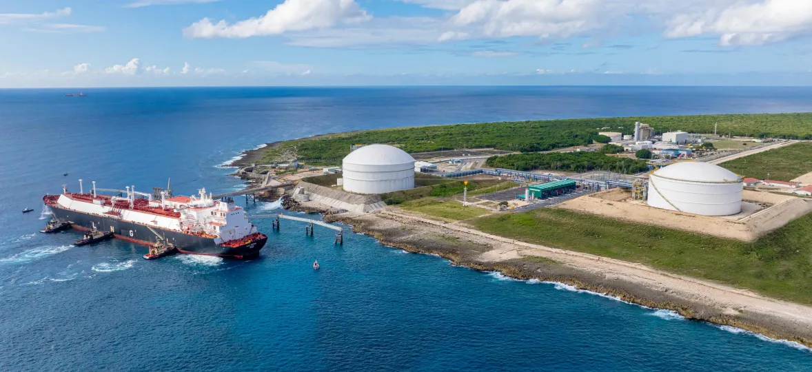 Aerial view of a coastal industrial area with two large white storage tanks near the shoreline. A large ship is docked at the pier, surrounded by clear blue water. The background shows green vegetation and a blue sky with scattered clouds.