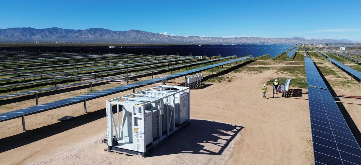 A large solar farm with rows of solar panels extending into the distance under a clear blue sky. In the foreground, there is a white equipment container on a dirt path. Two workers wearing yellow vests are near a set of panels, inspecting or working on them. Mountains are visible on the horizon.