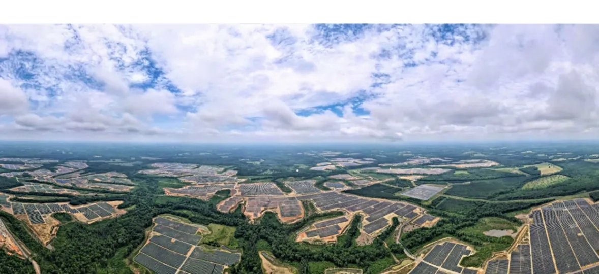 Aerial view of a large solar farm with numerous solar panels arranged in rectangular sections, surrounded by green forests and fields under a partly cloudy sky.