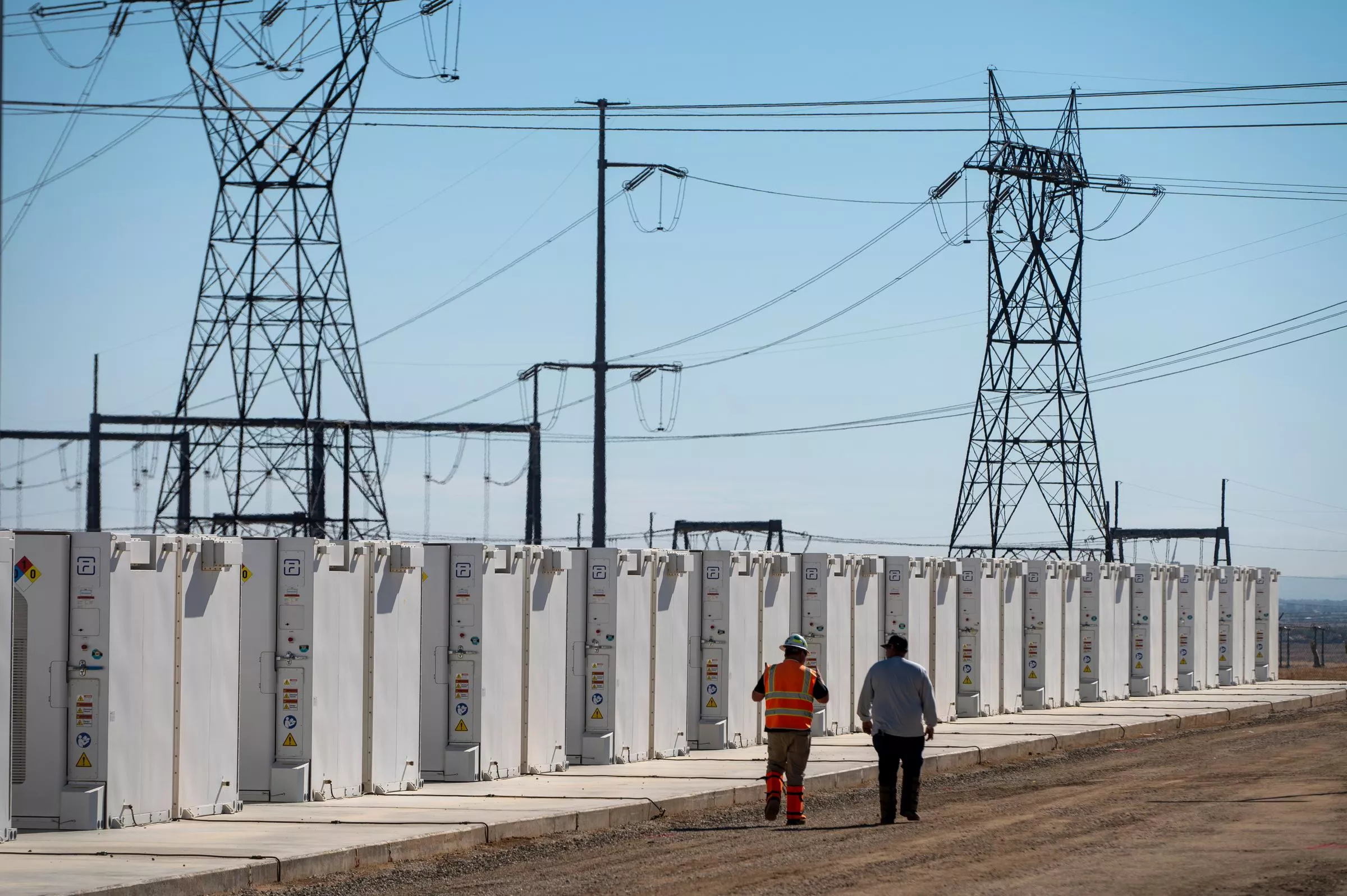 Two people walk along a row of large white battery storage units in an industrial area. High-voltage power lines and towers are visible in the background under a clear blue sky.
