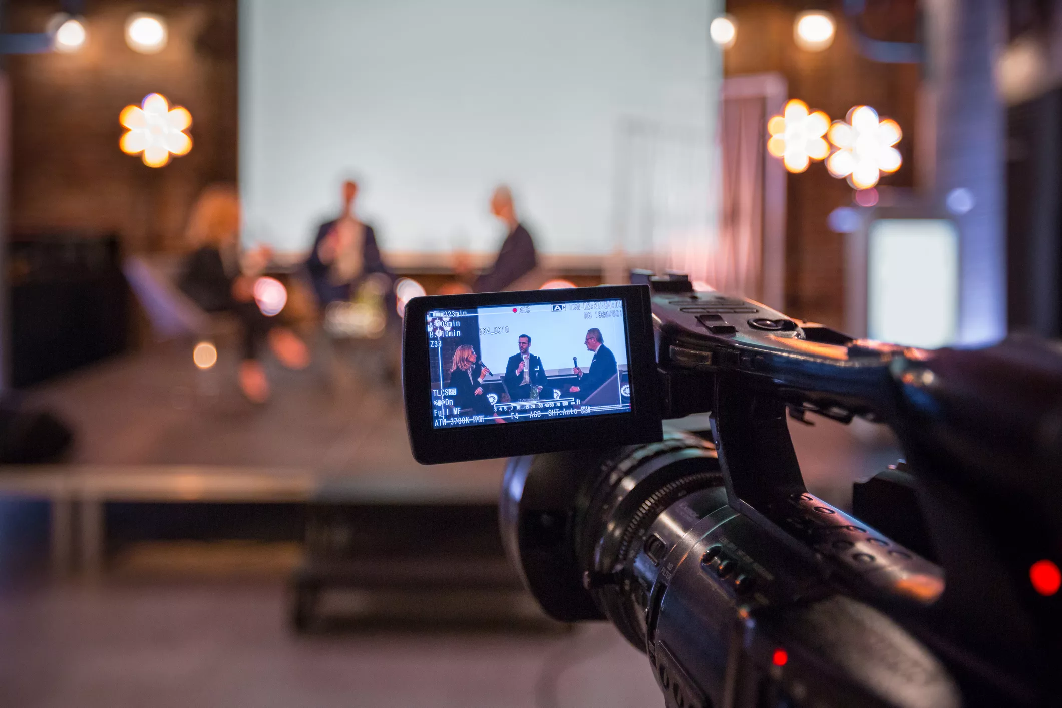 A video camera is focused on a panel discussion with three blurred individuals seated on a stage. The camera screen displays a clear image of the panelists talking. The background is softly lit with decorative lights.