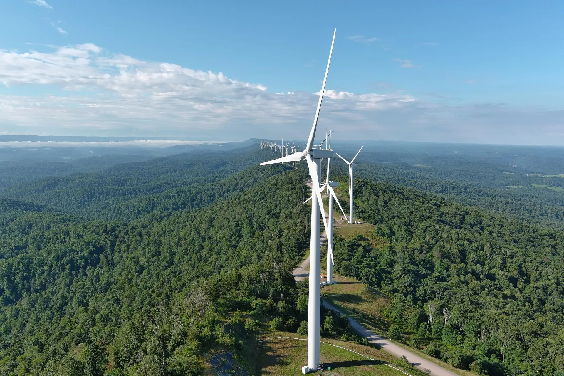 Aerial view of several wind turbines on a lush green hilltop, with a vast forested landscape and blue sky in the background.