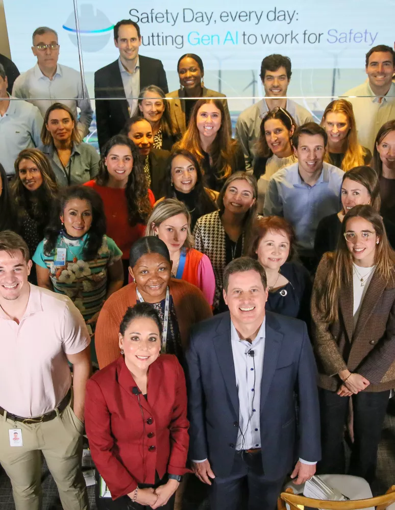 A large group of diverse people standing together in an office setting, smiling at the camera. A screen in the background displays the text: 'Safety Day, every day: Putting Gen AI to work for Safety.'