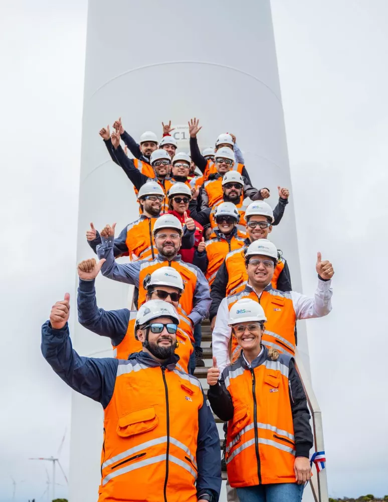 A group of people wearing orange safety vests and helmets stand on a staircase in front of a wind turbine, giving thumbs up. The sky is overcast and other wind turbines are visible in the background.