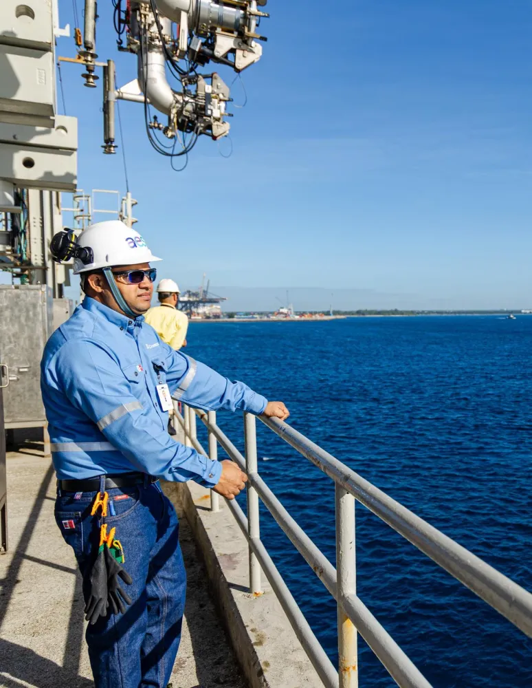 A worker wearing a blue uniform and white hard hat stands on a platform overlooking a large body of water. The worker is holding onto a metal railing. In the background, there is a small boat on the water and a distant shoreline with industrial structures.