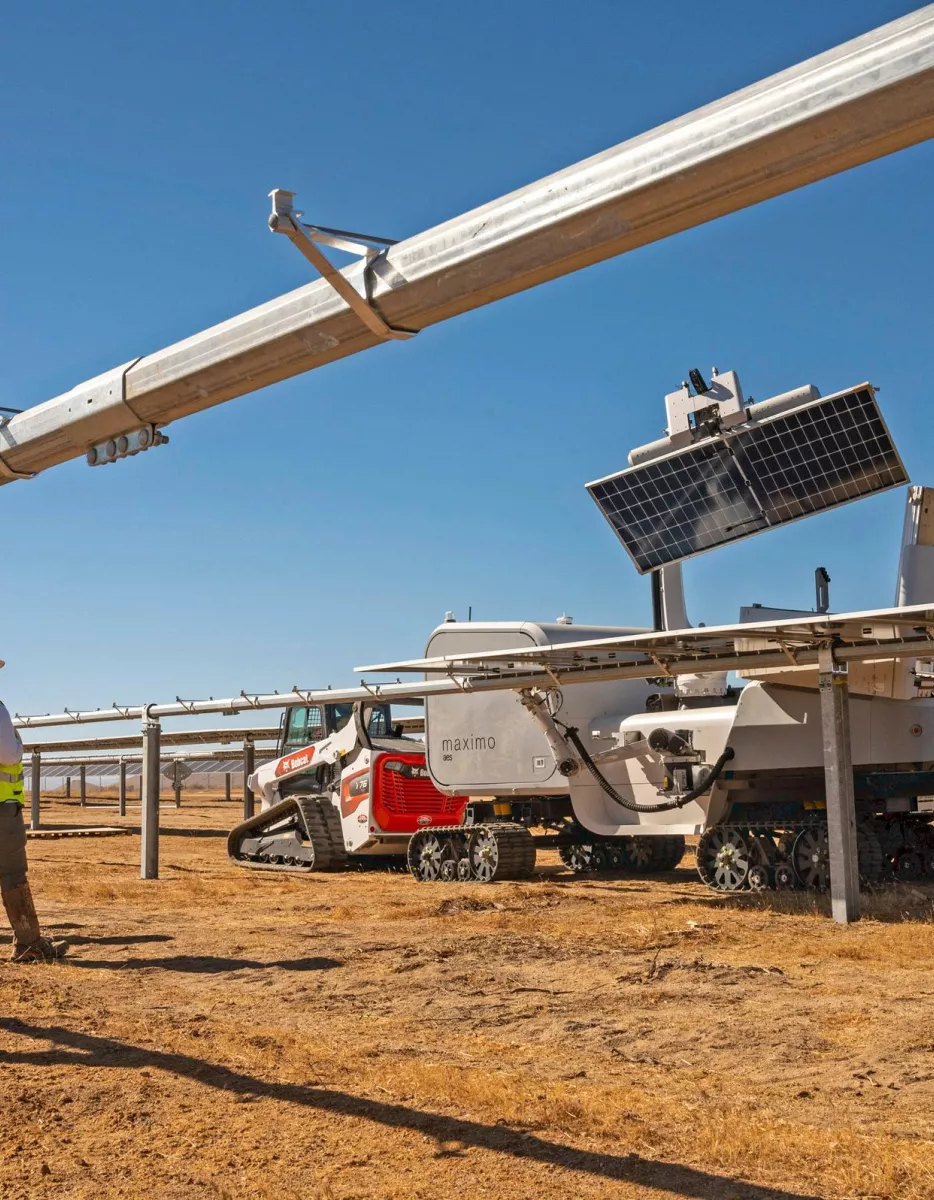 Three workers in safety gear stand beside a solar panel installation in a dry, open field. A robotic machine with tracks is positioned under the panels, likely for maintenance or cleaning. The sky is clear and blue.
