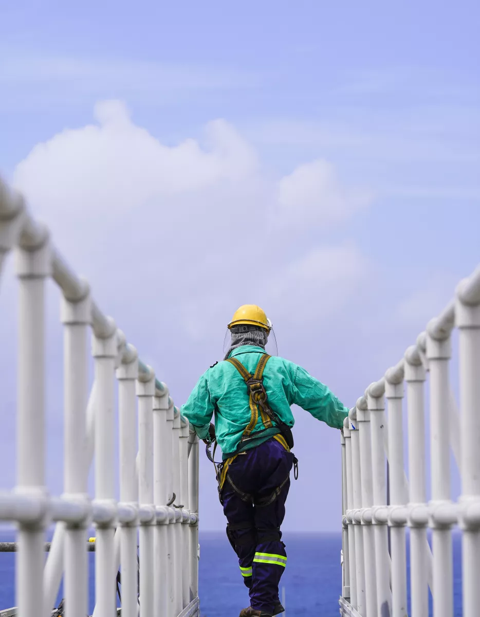 A worker wearing safety gear, including a yellow helmet and green jacket, walks along a metal walkway with railings, set against a backdrop of clear blue sky and ocean.