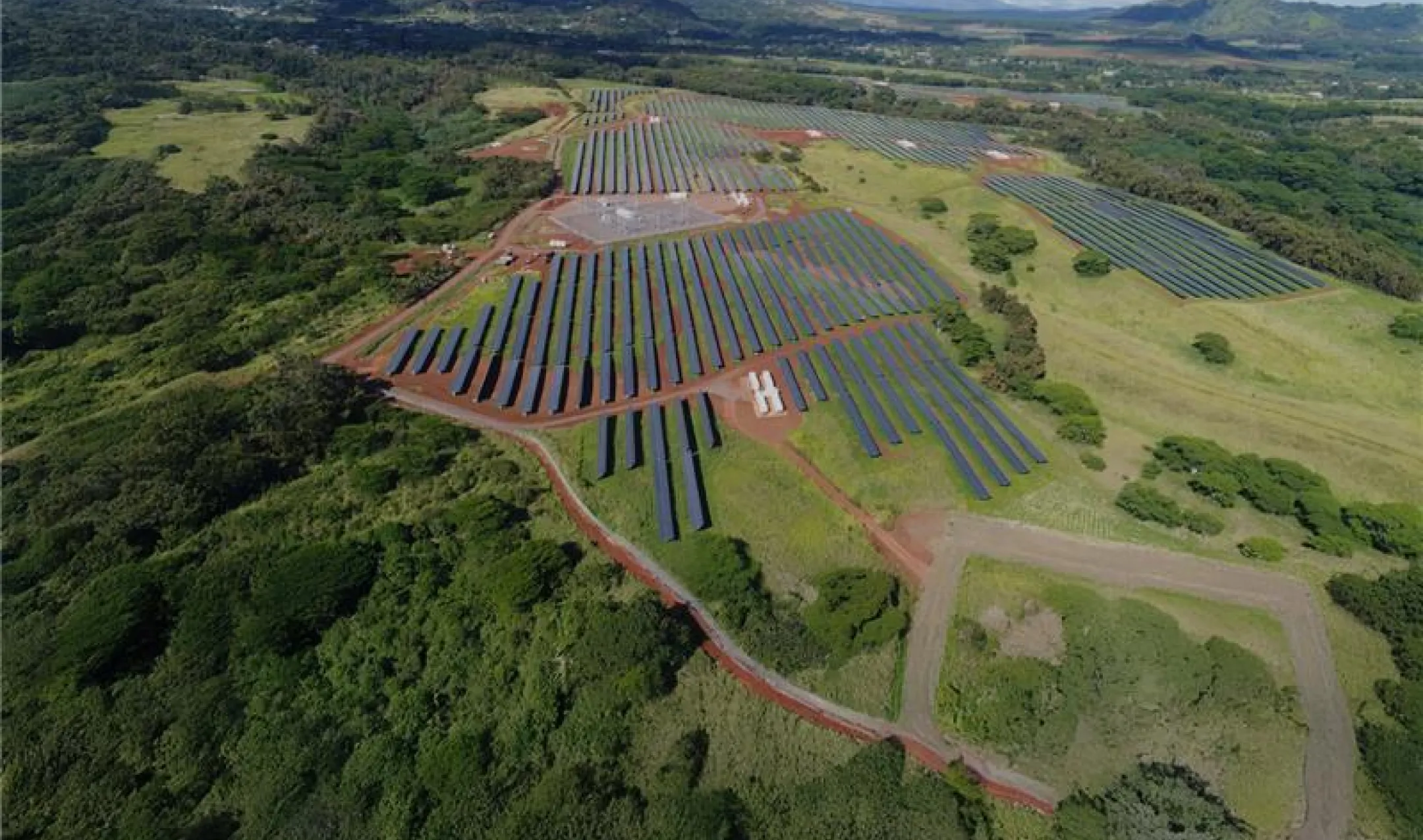 Aerial view of a large solar farm with multiple rows of solar panels set in a green landscape surrounded by hills and trees under a partly cloudy sky.