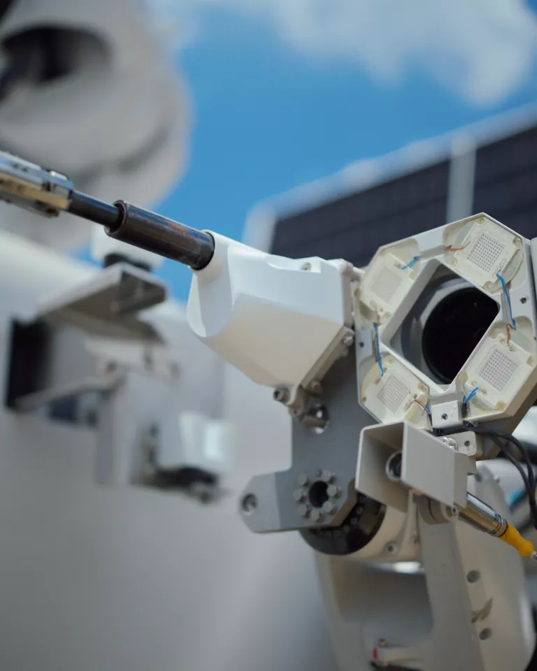 Close-up of a mechanical robotic arm with a cylindrical tool attached. The arm features various components, wires, and connectors, set against a blurred background with a solar panel and blue sky.