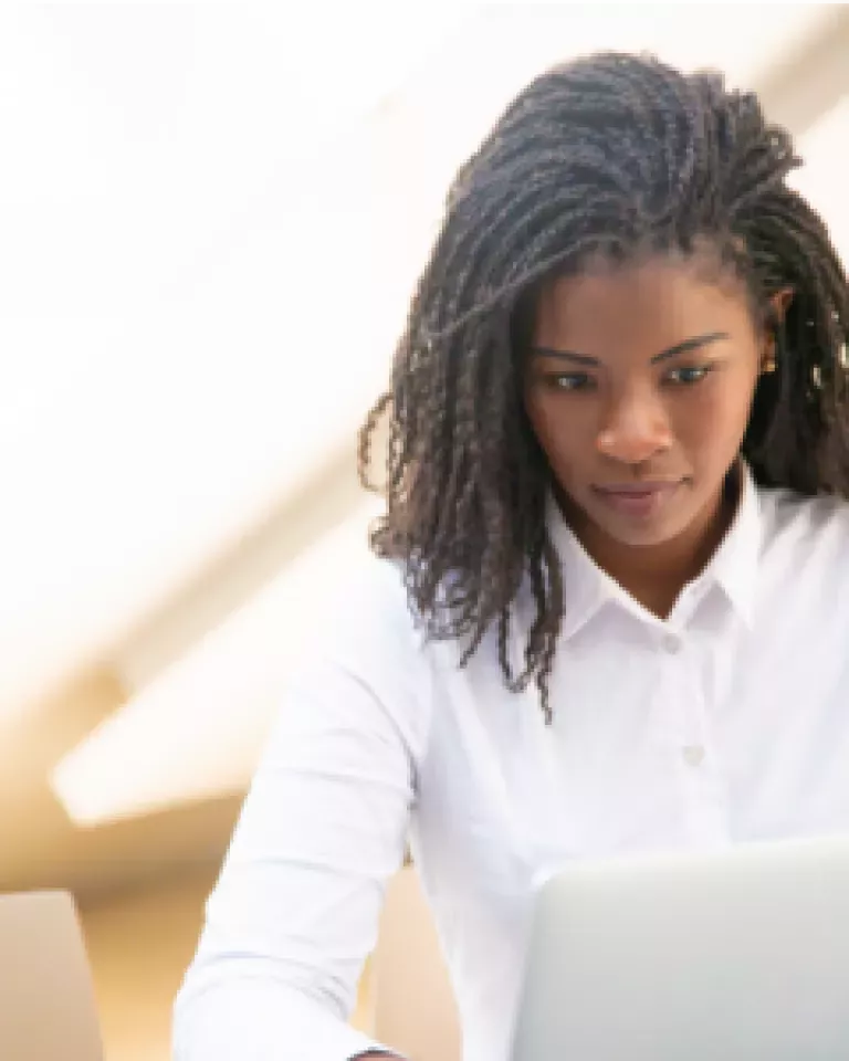 A person with braided hair wearing a white shirt is looking at a laptop screen with a focused expression.