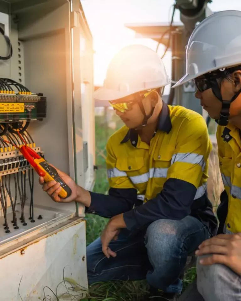 Two workers in yellow safety jackets and helmets examine electrical equipment in an outdoor setting. One holds a multimeter to test the wires inside an open panel, while the other observes. The sun is setting in the background.