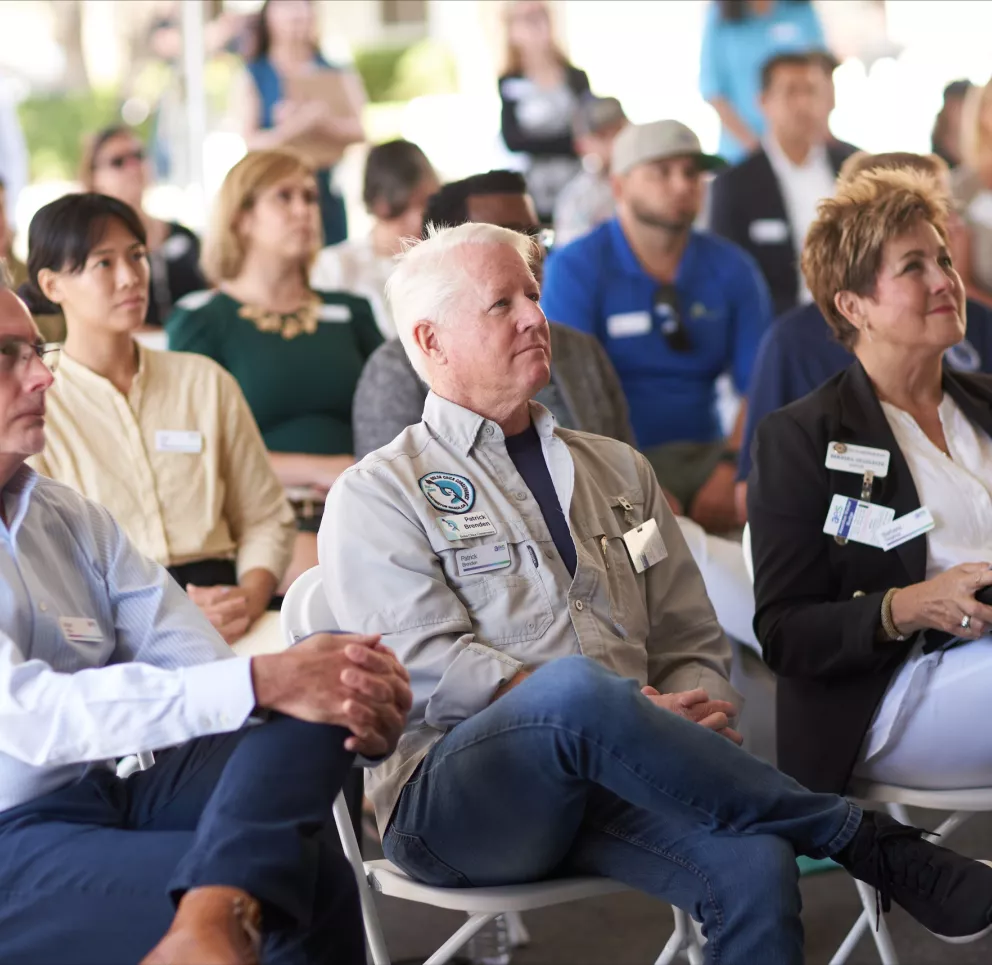 A group of people sitting on white folding chairs attentively listening to a presentation. They are in an outdoor setting, with some wearing name tags and business casual attire. The focus is on three individuals in the front row, with others blurred in the background.