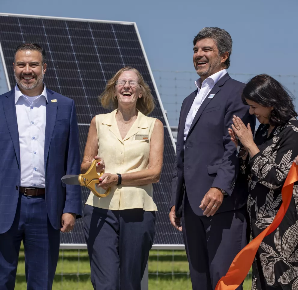 Group of people smiling at a ribbon-cutting ceremony in front of a solar panel. A woman holds large scissors, and others hold an orange ribbon.