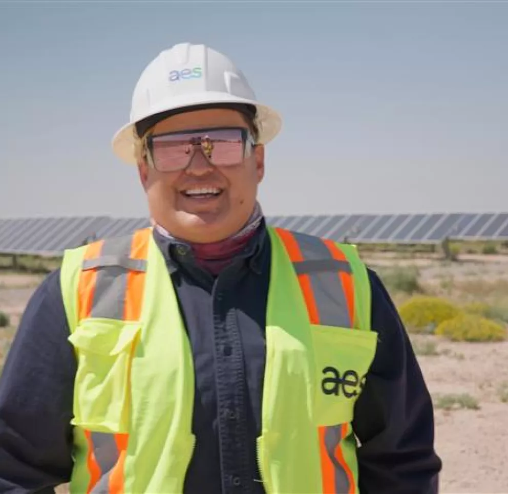 A person wearing a white hard hat and reflective safety vest stands in front of a solar panel field. The person is smiling and wearing sunglasses, with a clear sky in the background.