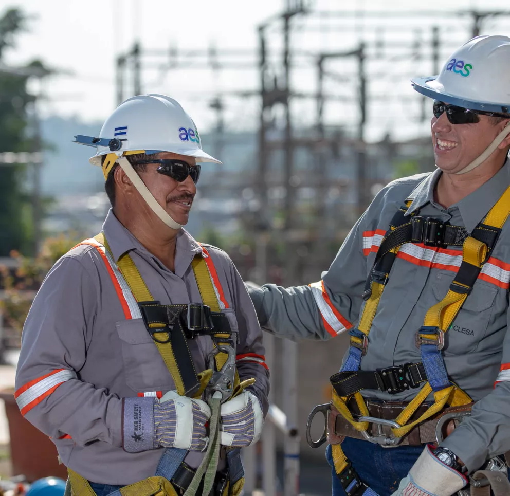 Two construction workers wearing safety helmets and harnesses smile and interact at a work site. Both are wearing gray uniforms with orange stripes. One worker has a hand on the other's shoulder. Electrical equipment and trees are visible in the background.