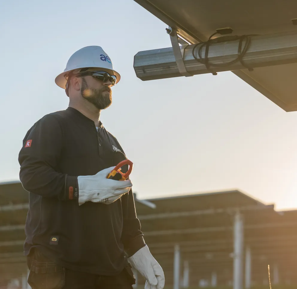 A worker wearing a hard hat and sunglasses stands next to a solar panel, holding a tool. The sun is setting in the background, casting a warm glow over the scene.