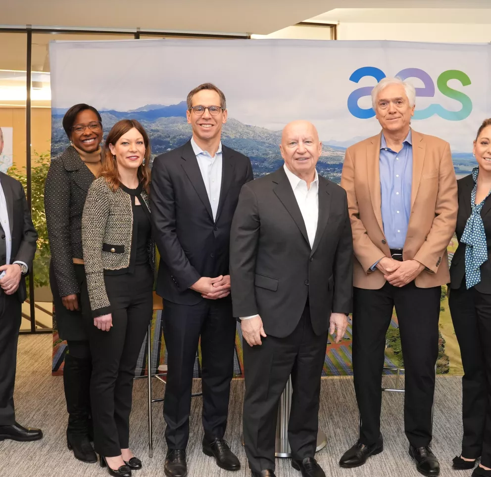 A group of nine people stand in a row, smiling, in front of a backdrop with the AES logo. They are dressed in business attire, including suits and blazers. The setting appears to be an office environment with a modern design.