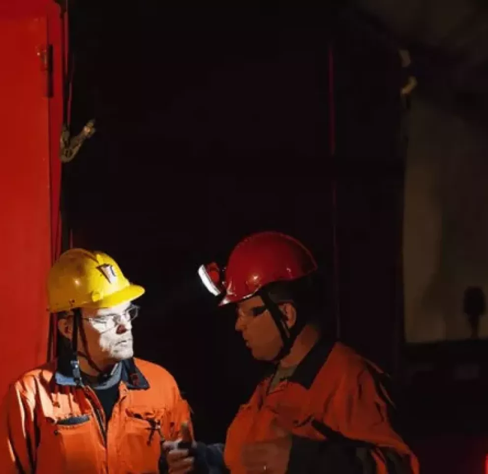 Two miners wearing orange uniforms and helmets stand in a dimly lit mining tunnel. One helmet is yellow, and the other is red with a headlamp. They are engaged in conversation next to a large red door marked with 'Mi'.