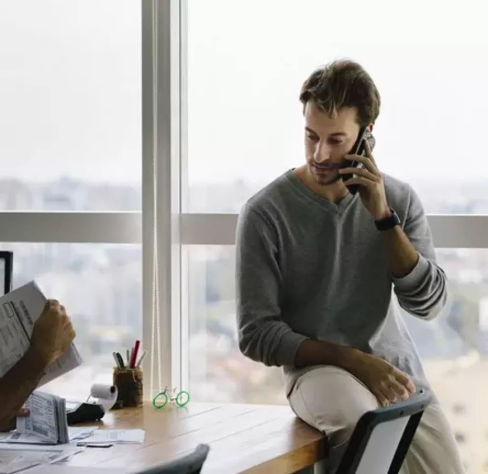 Two men in an office with large windows. One is sitting at a desk reviewing documents, while the other is sitting on the windowsill talking on a phone. The background shows a cityscape view.
