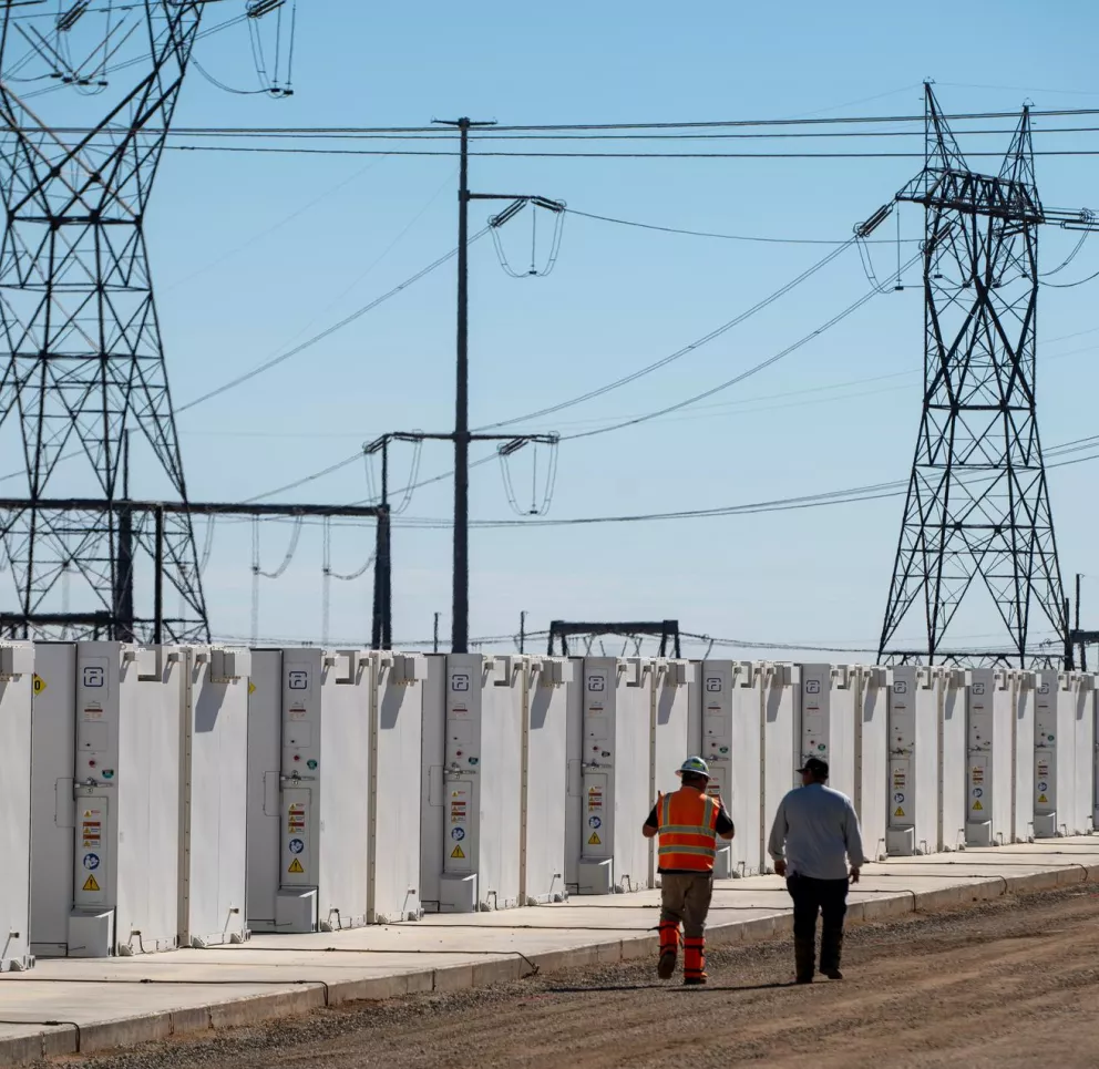 Two workers walk alongside a row of white industrial battery storage units in a power facility. High voltage transmission towers and power lines are visible in the background under a clear blue sky.