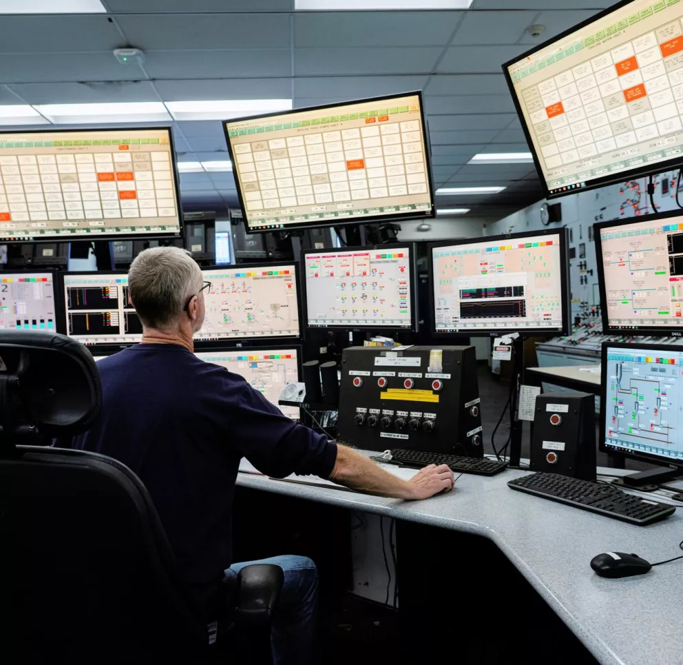 A person sits at a control room desk surrounded by multiple computer monitors displaying complex data and diagrams. The screens are arranged in a semicircle, showing various technical interfaces and charts. The person is focused on the screens, suggesting a monitoring or management task in a high-tech environment.