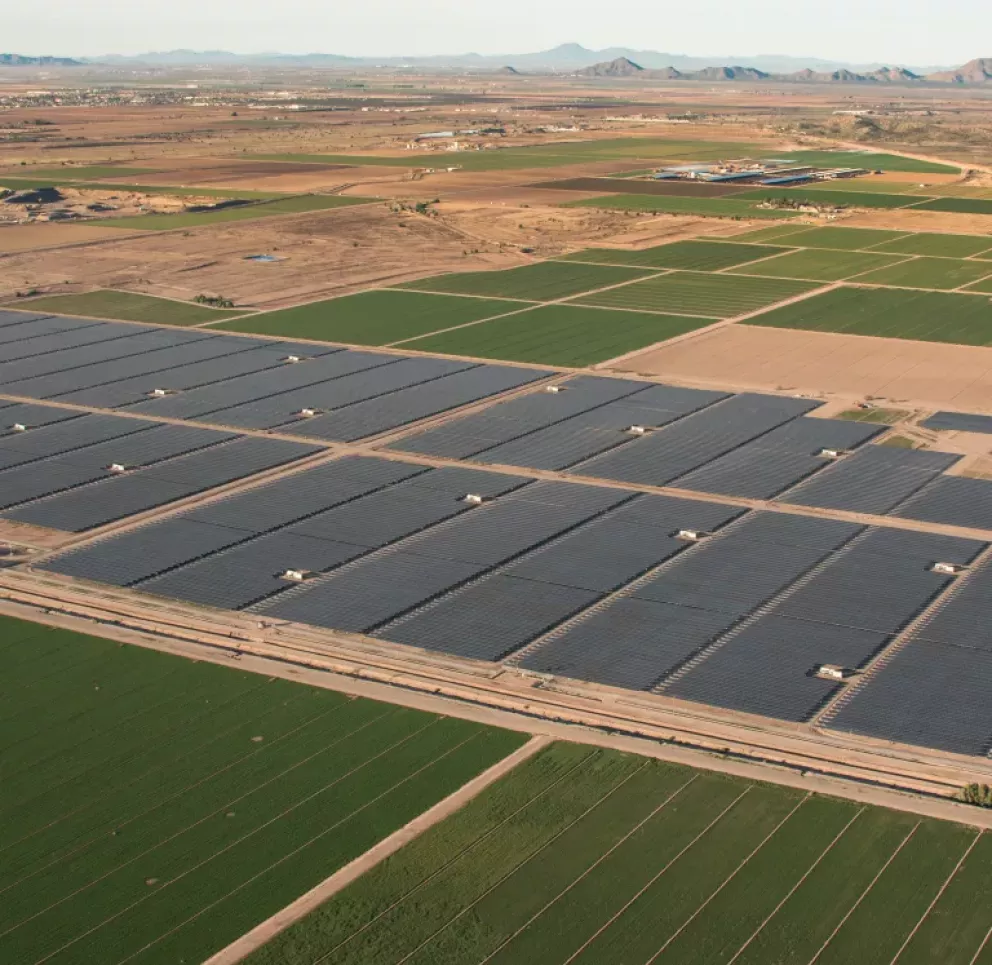 Aerial view of a large solar farm with rows of solar panels adjacent to green agricultural fields. The landscape extends to distant mountains under a clear sky.
