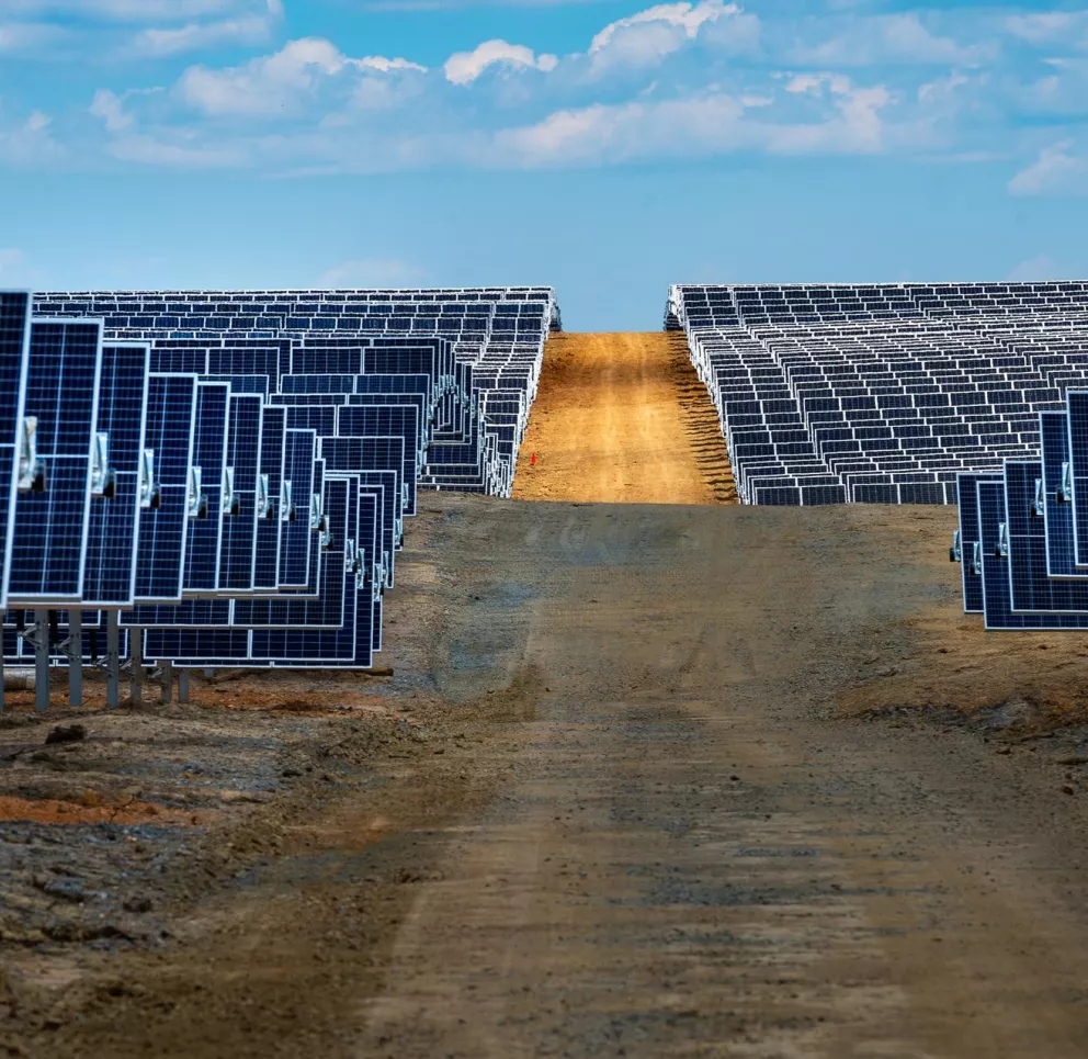 A large solar farm with rows of solar panels on both sides of a dirt path, extending into the distance under a partly cloudy sky.