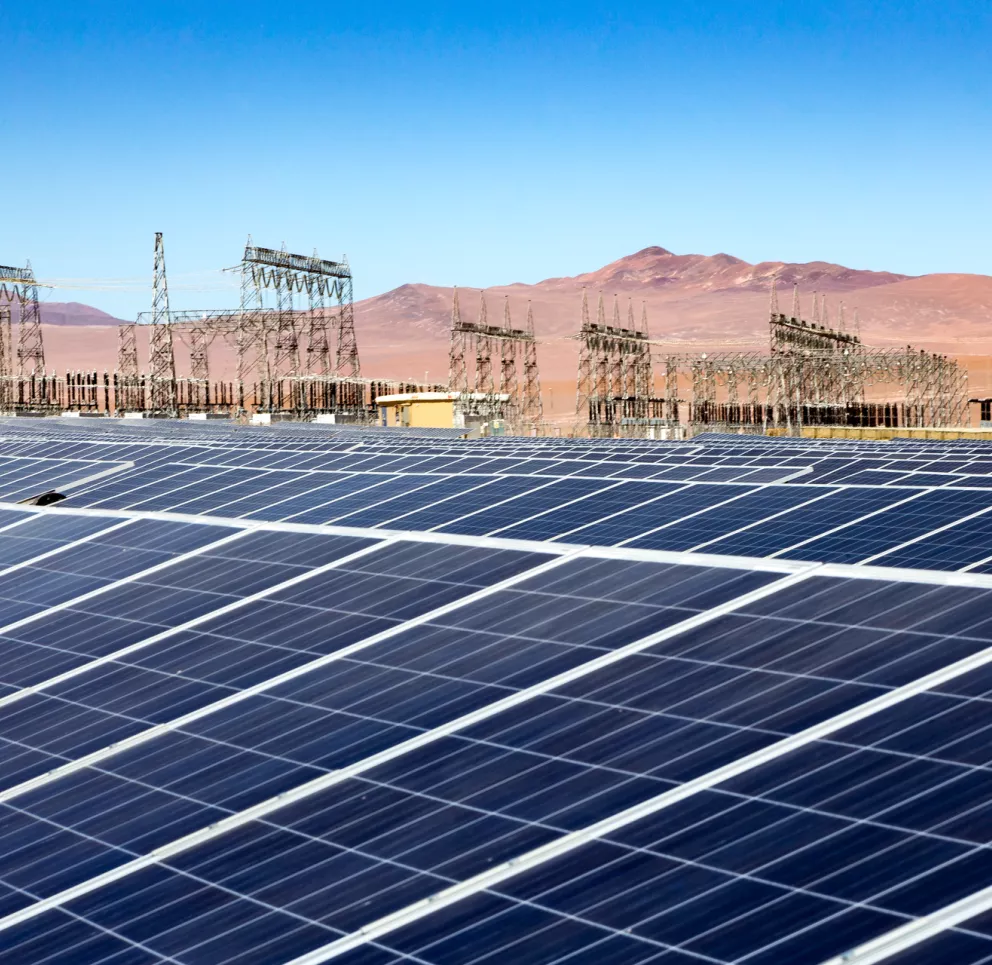 A large solar panel array in the foreground with an electrical substation and transmission towers in the background. The landscape is arid with distant mountains under a clear blue sky.