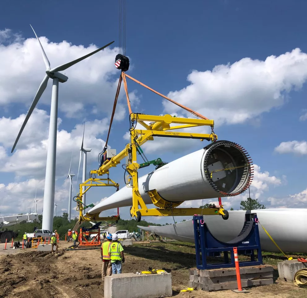 Construction workers in safety vests guide a large wind turbine blade being lifted by a crane. Several standing wind turbines are visible in the background under a blue sky with clouds.
