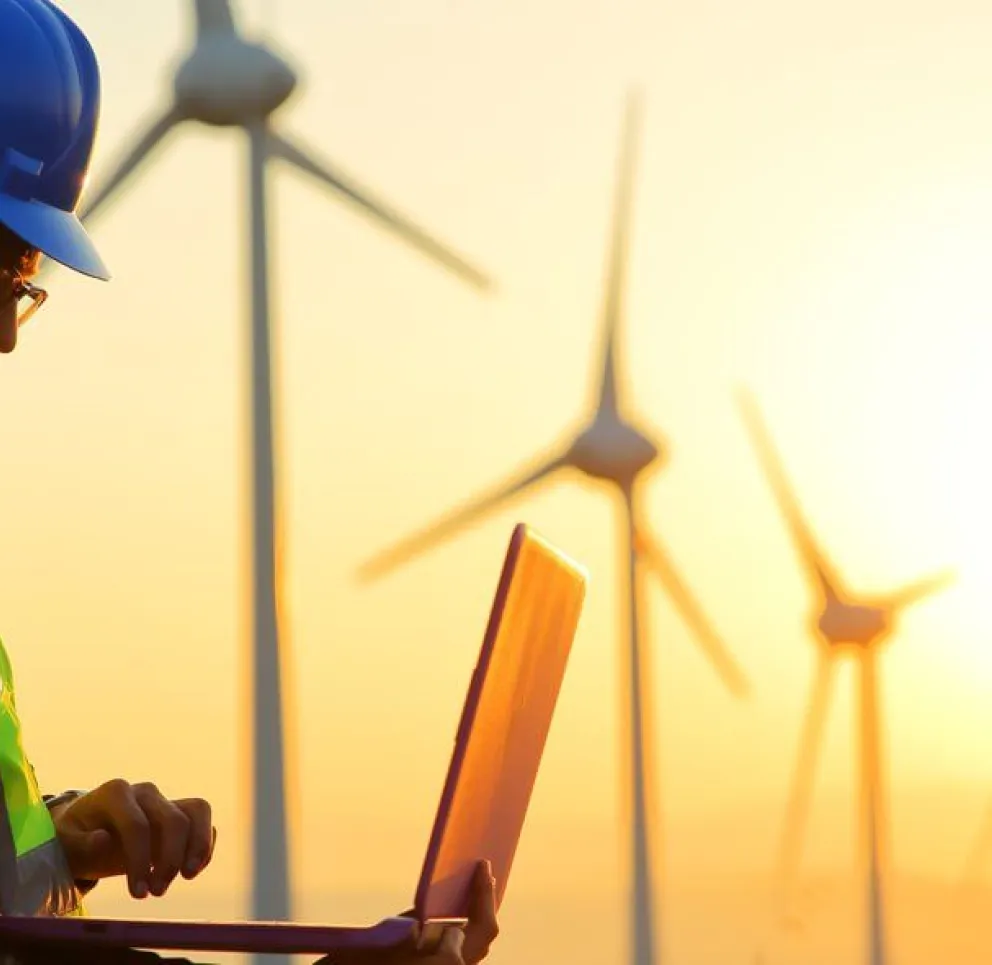 Two workers wearing hard hats and reflective vests stand outdoors with a laptop, analyzing data in front of wind turbines at sunset.