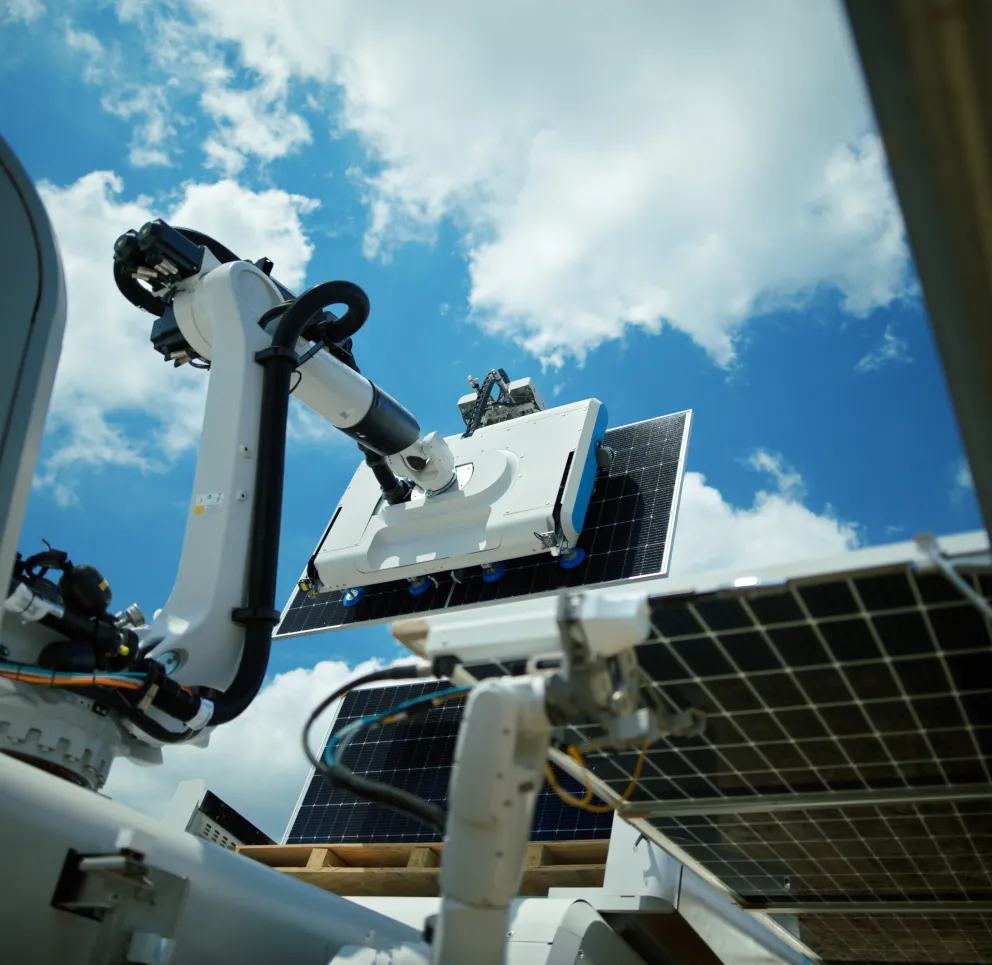 A robotic arm with a cleaning device is positioned over a solar panel under a bright blue sky with scattered clouds.