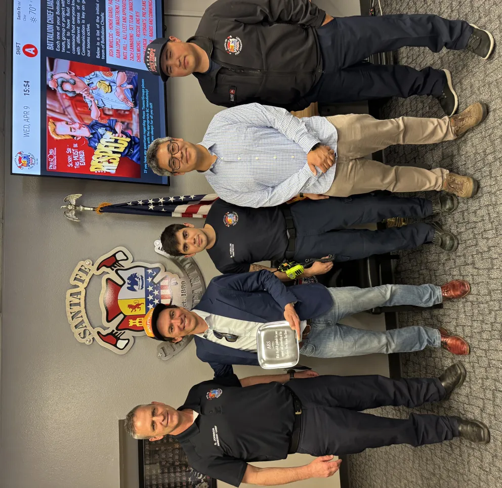 Five men stand in a row in front of a Santa Fe fire department emblem on a wall. One man holds a plaque. They are dressed in a mix of casual and uniform attire. An American flag and a digital screen displaying information are visible in the background.