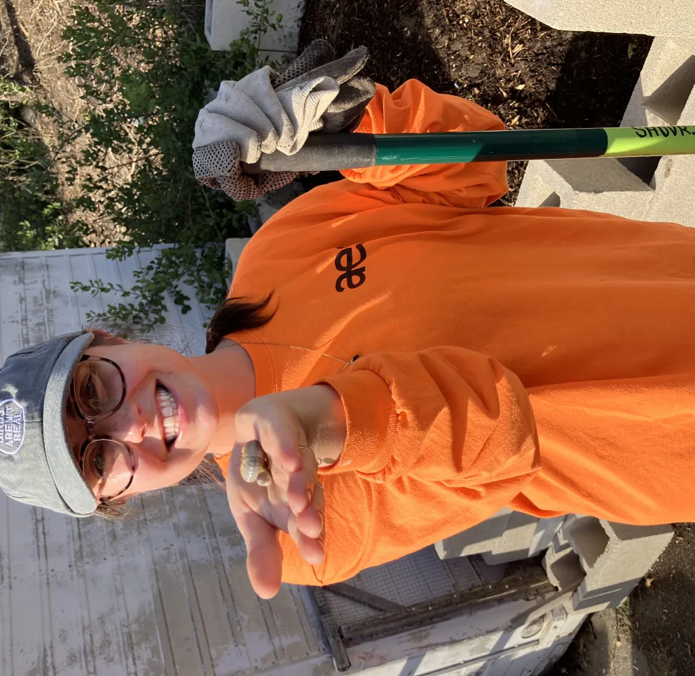 A person wearing glasses, a gray cap, and an orange shirt smiles while holding a small snail in their hand. They are standing outdoors near a pile of cinder blocks and holding a gardening tool with gloves draped over it.