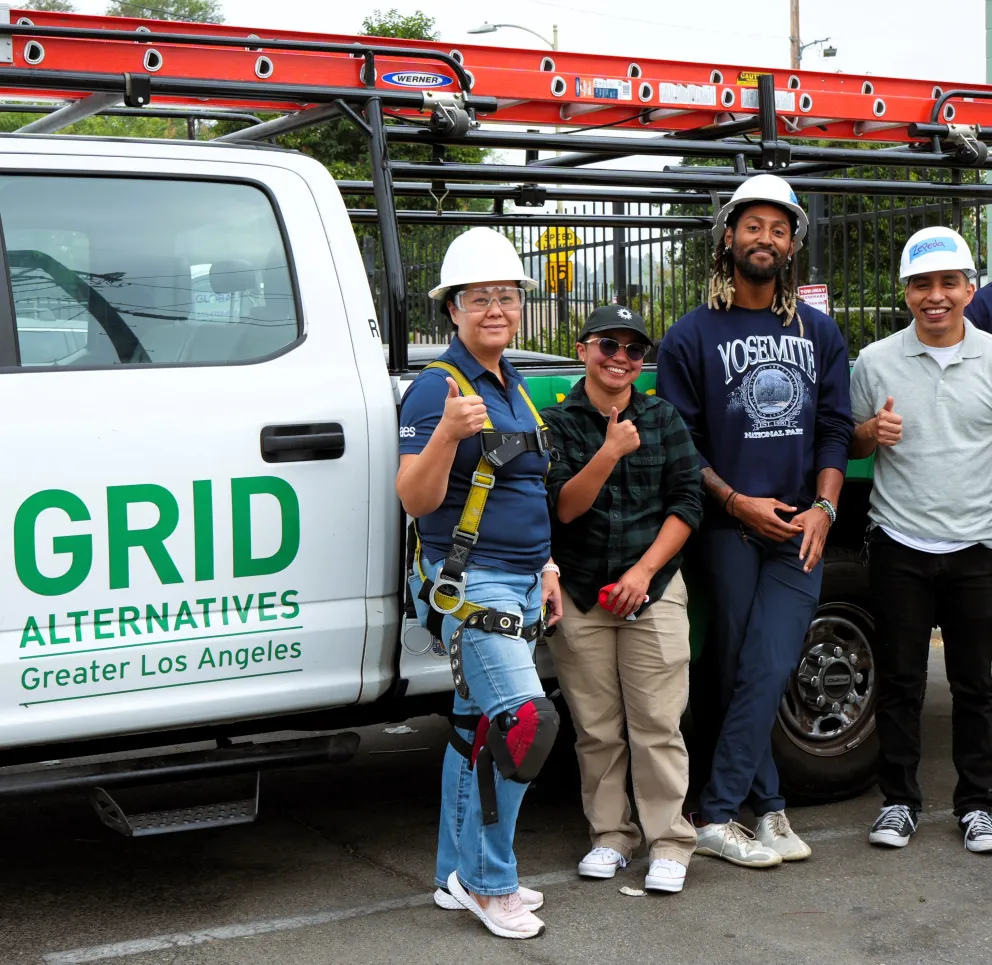 A group of seven people standing next to a white truck with 'GRID Alternatives Greater Los Angeles' logo. They are wearing casual clothes and some have hard hats. They are smiling and giving thumbs up.