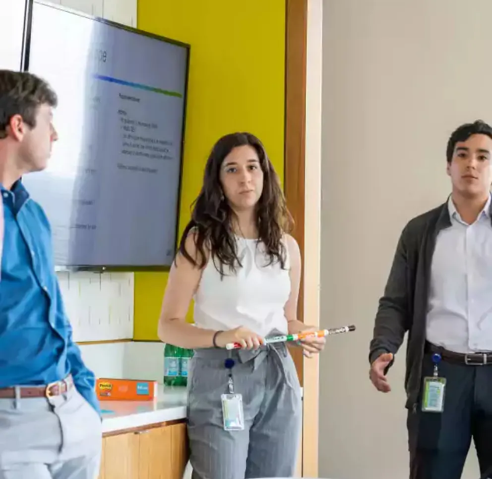 Three young professionals standing in a modern office, one woman holding a marker and presenting in front of a screen with a presentation slide, while two men listen attentively.
