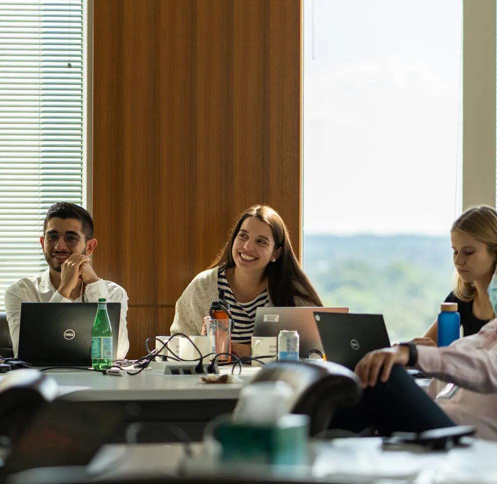 A group of young adults sitting at a conference table with laptops, water bottles, and notebooks, attentively listening and smiling during a meeting in a well-lit office with large windows.