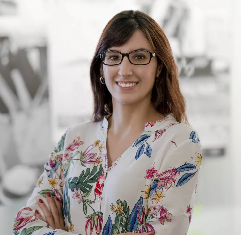 Woman with long brown hair and glasses smiling confidently, wearing a white blouse with colorful floral patterns, standing with arms crossed in a bright, blurred indoor setting.