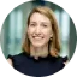 A person with shoulder-length hair smiling, wearing a dark top. The background is softly blurred, suggesting an indoor setting.