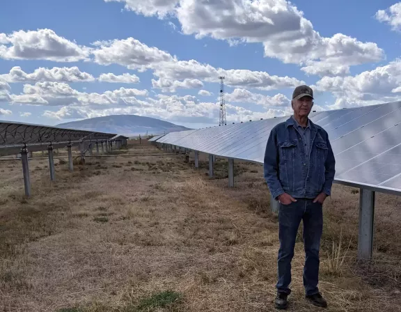 A person in a denim jacket and cap stands in a dry field next to rows of solar panels under a partly cloudy sky.