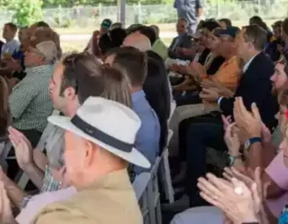 A large group of people seated under a tent, clapping their hands. The audience is diverse, with individuals of various ages and attire, including hats and casual clothing. The setting appears to be an outdoor event with trees visible in the background.