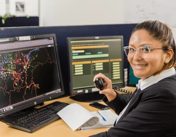 A person in an office setting is smiling while holding a phone handset. They are seated at a desk with two computer monitors displaying complex data and maps. A notebook with a pen is open on the desk.