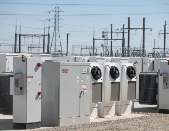 A row of large electrical transformers and equipment in an outdoor substation. The equipment is marked with '4200 kVA' and features cooling fans. In the background, there are power lines and transmission towers under a clear blue sky.