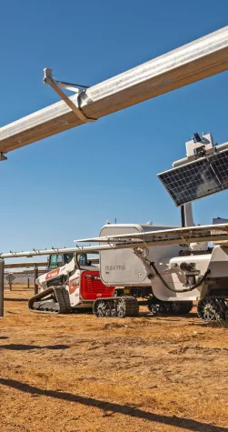 Three workers in safety gear stand beside a solar panel installation in a dry, open field. A robotic machine with tracks is positioned under the panels, likely for maintenance or cleaning. The sky is clear and blue.