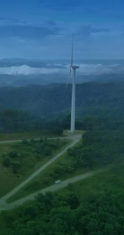 Aerial view of a lush green landscape with multiple wind turbines scattered across the hills. A winding road leads through the forested area, with a single vehicle visible on it. The sky is clear with a few clouds on the horizon.
